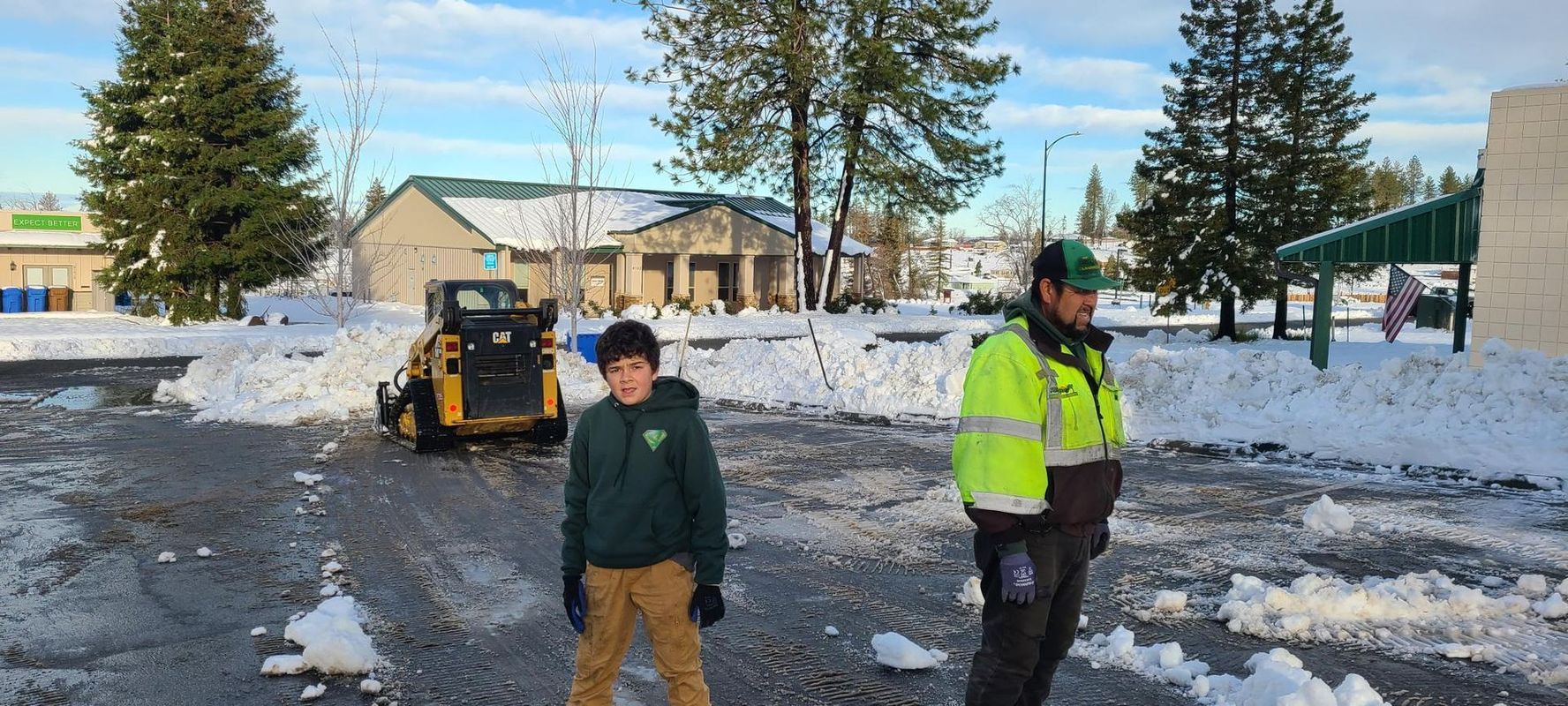 A person and a worker stand in a snowy parking lot as a small tractor clears snow from it. A building is in the background.