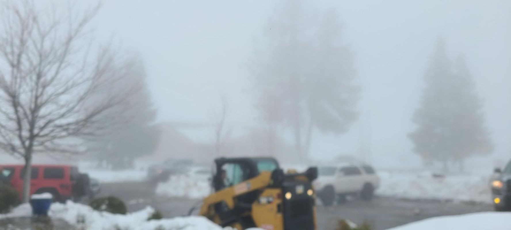 A yellow skid steer clears snow in a snowy, foggy residential area, with cars and bare trees.