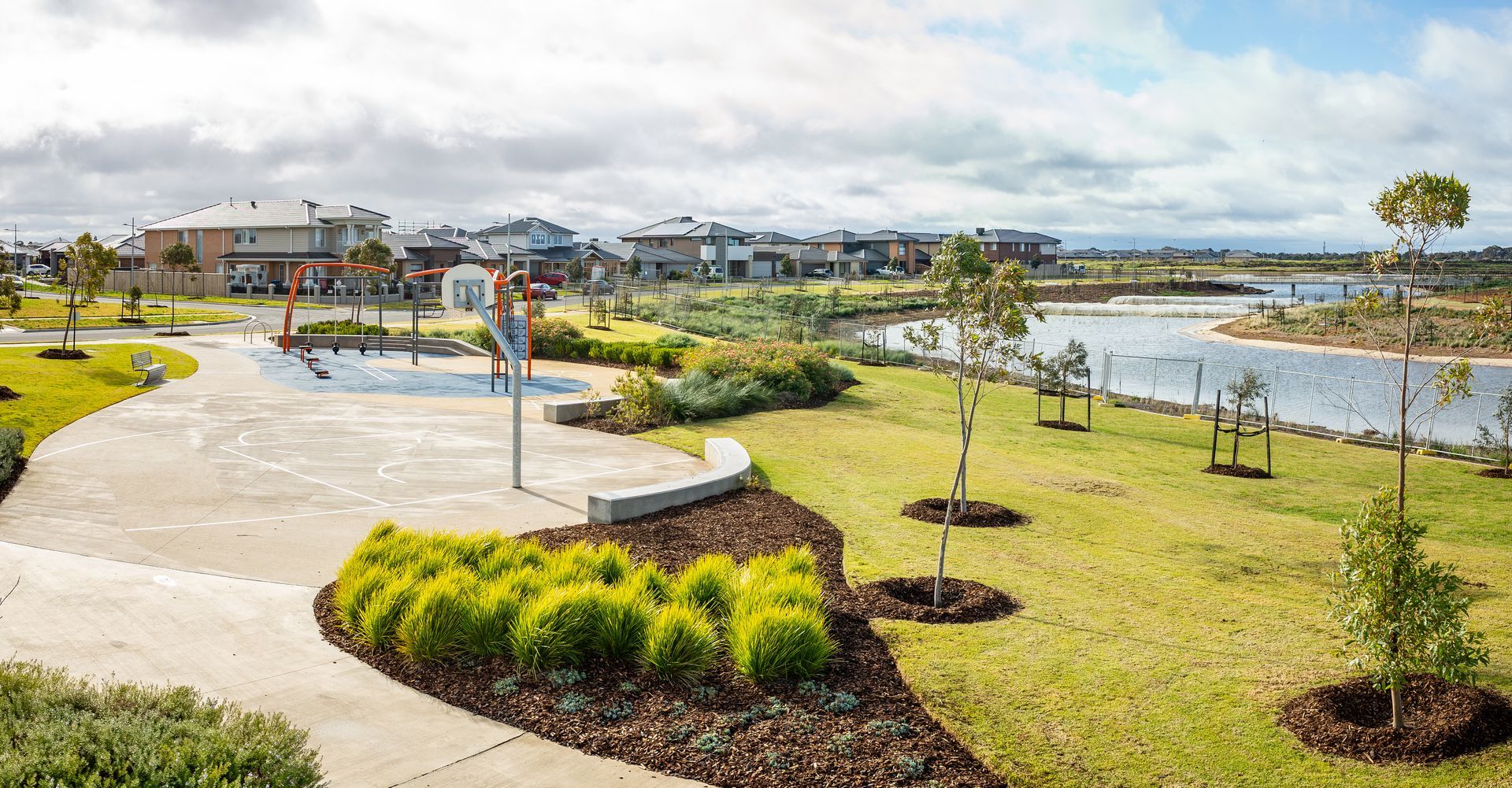 Park with basketball court, houses, water, and green space under cloudy sky.