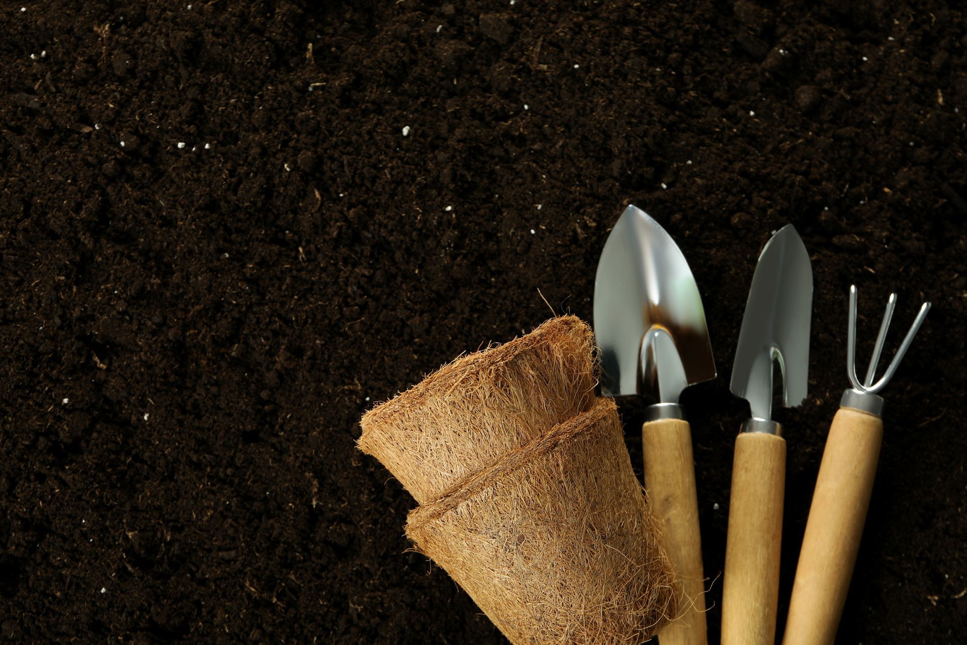 Gardening tools, including a trowel, spade, and hand rake, next to peat pots in dark soil.