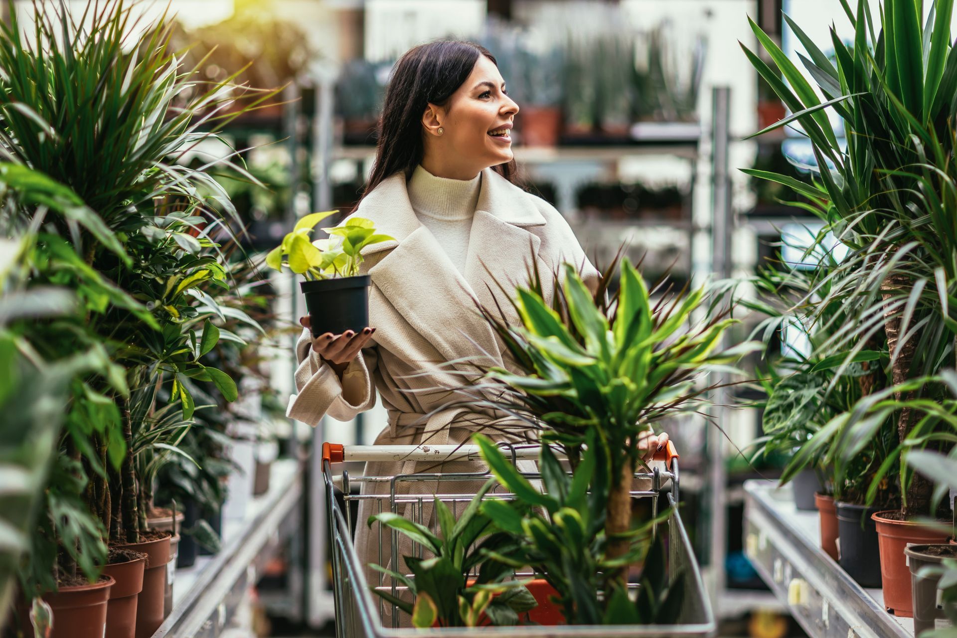 Woman shopping for plants in a garden center, holding a potted plant, smiling, pushing a cart.