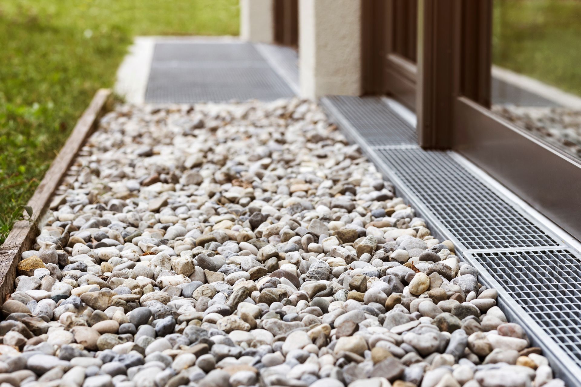 Gravel bed next to a building with a metal grate drain, brown window frame, and green lawn.