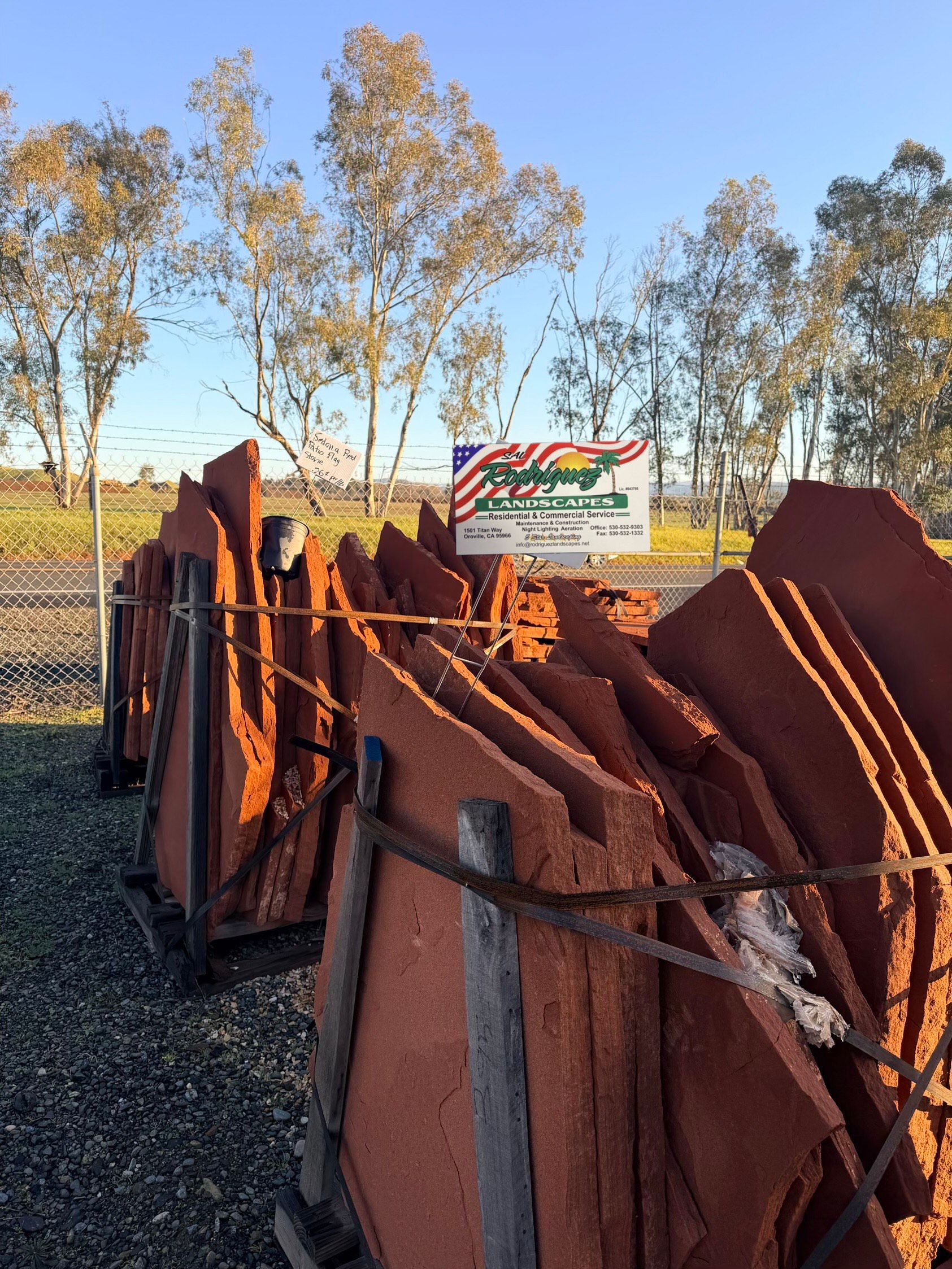 Stacks of reddish-brown roof tiles on pallets, with a sign in the background and trees under a blue sky.