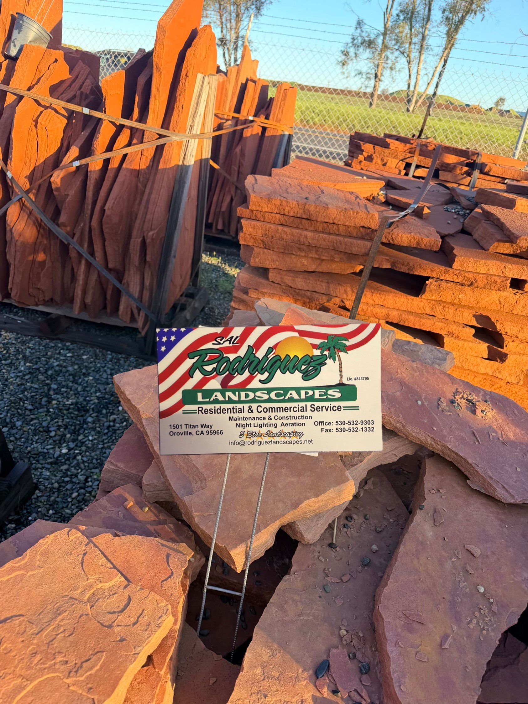 Piles of reddish-brown stone slabs with a sign for Tropical Landscapes USA.