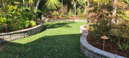 Curved stone retaining walls frame a green lawn, edged by lush plants and trees in a sunny garden.