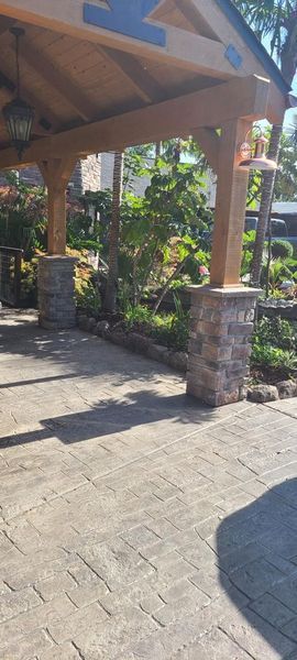 Wooden-beamed entryway with brick columns and paved walkway. Lush greenery in the background.