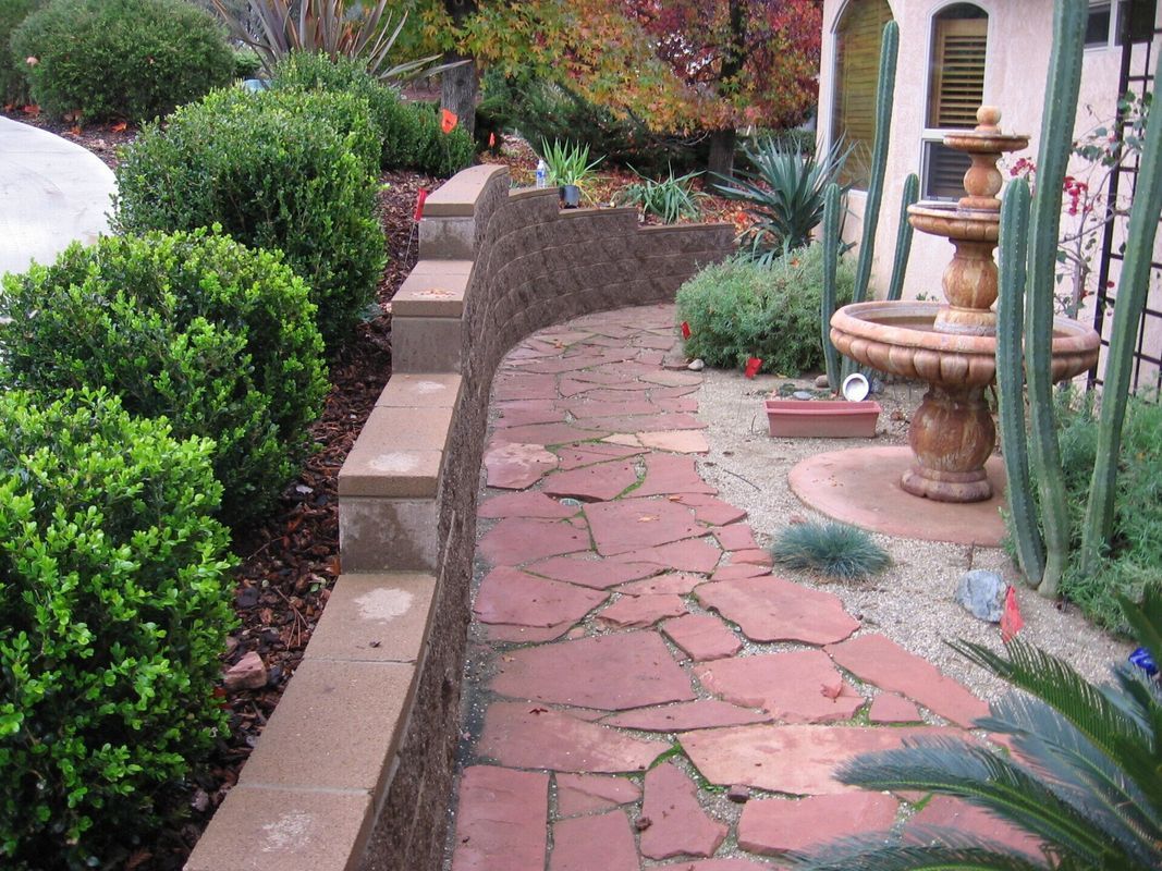 Red brick pathway curves through a garden with a fountain and shrubbery.