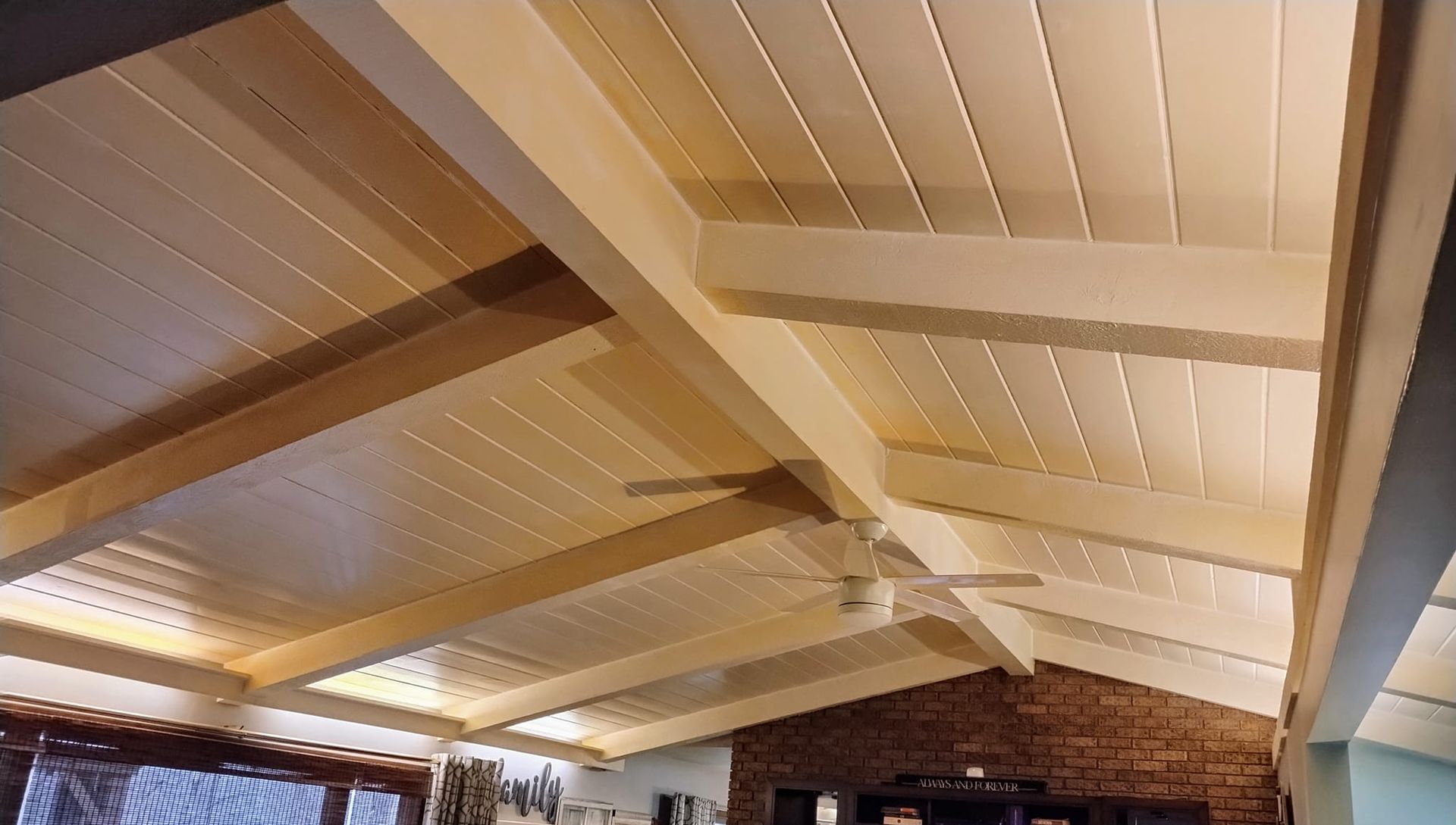 Beige wooden slatted ceiling with beams, a fan, and partial view of a brick wall.