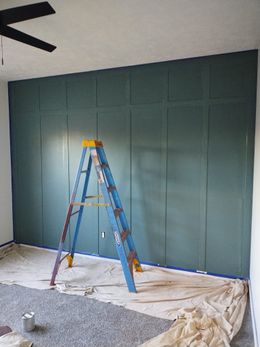 A blue ladder leans against a teal-paneled wall, with drop cloths and paint cans on the floor.