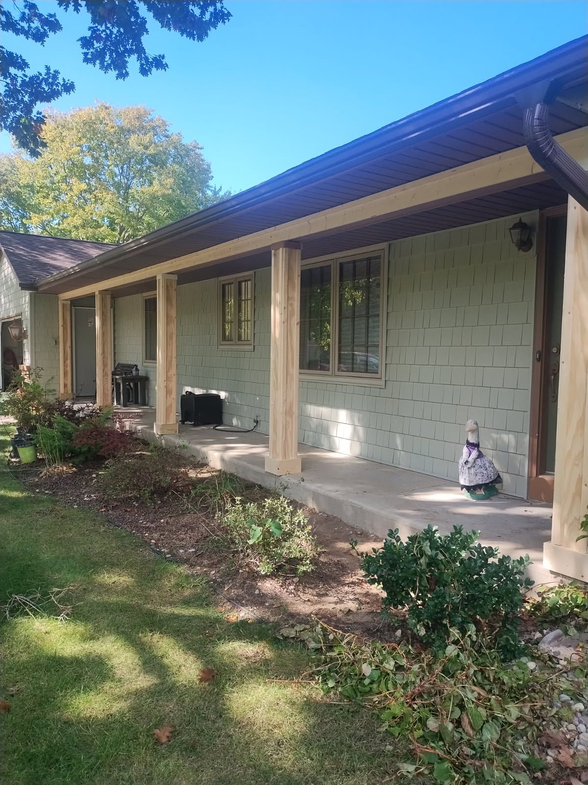 Exterior of a house with porch posts. Tan siding, brown trim, and lush greenery in front. Blue sky above.