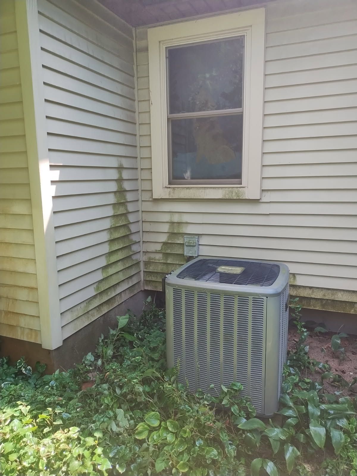Air conditioning unit near a house with weathered siding and a window.