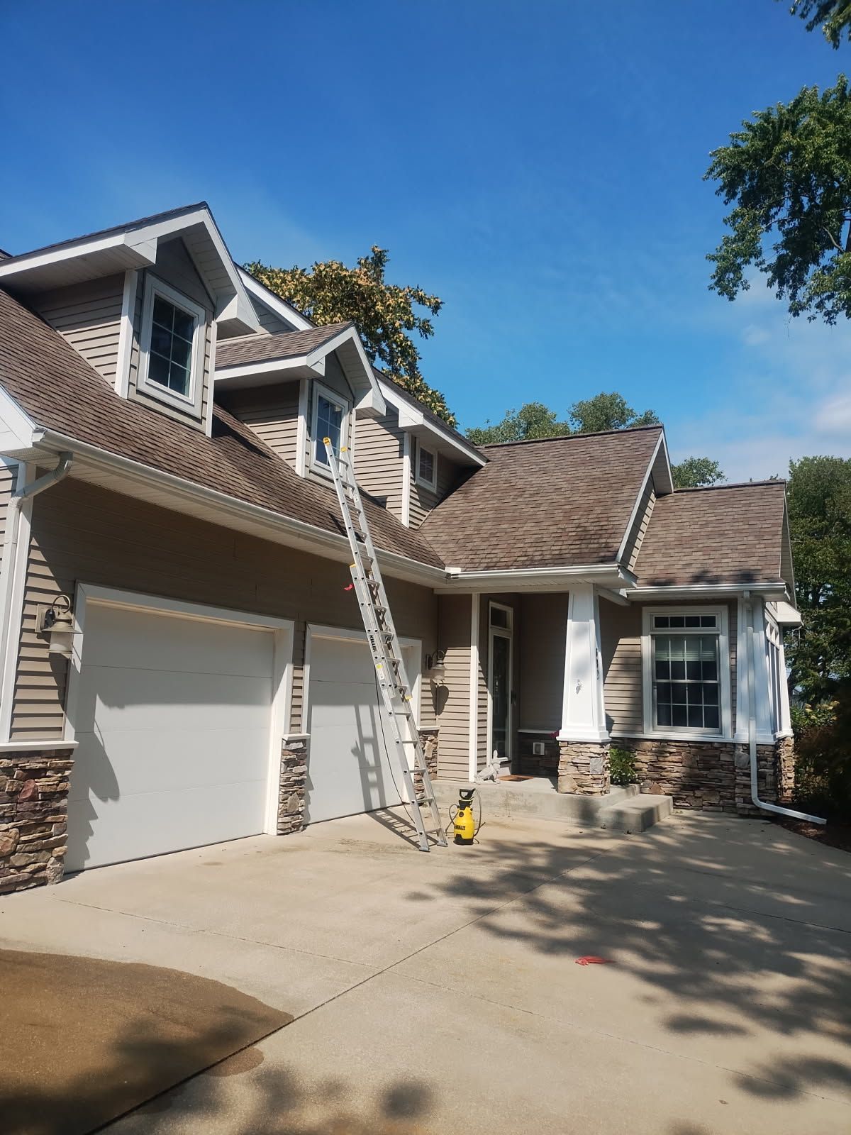 House with tan siding and stone accents, ladder leading to the roof, blue sky overhead.