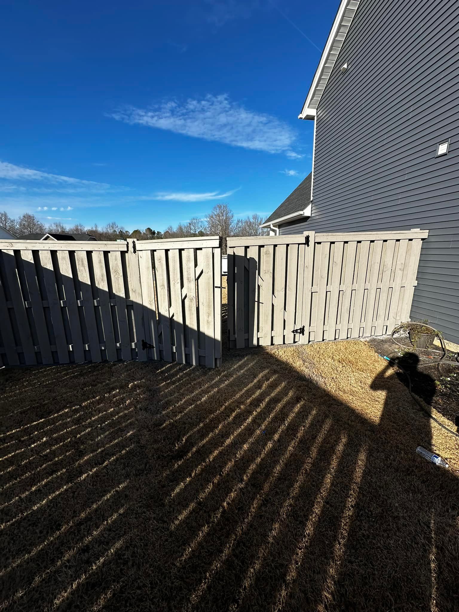 Fence and patterned house exterior with long shadows on gravel ground under a blue sky.