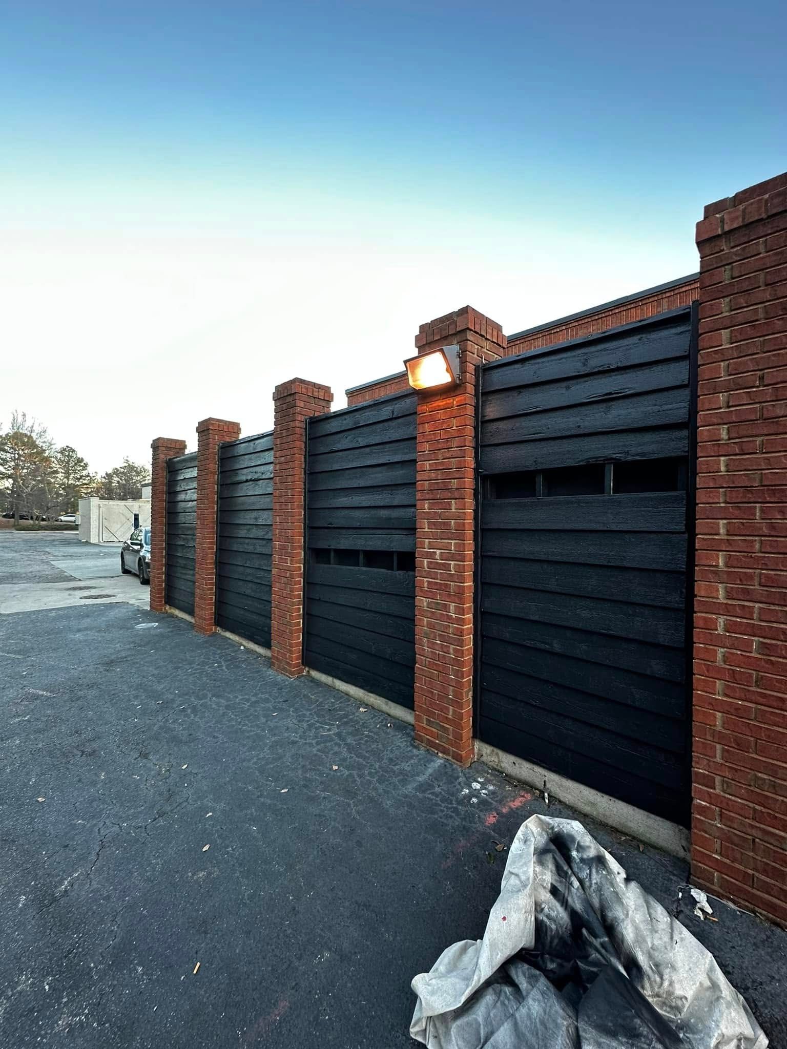 Black wooden fence with brick pillars and a light, set on a gravel surface.