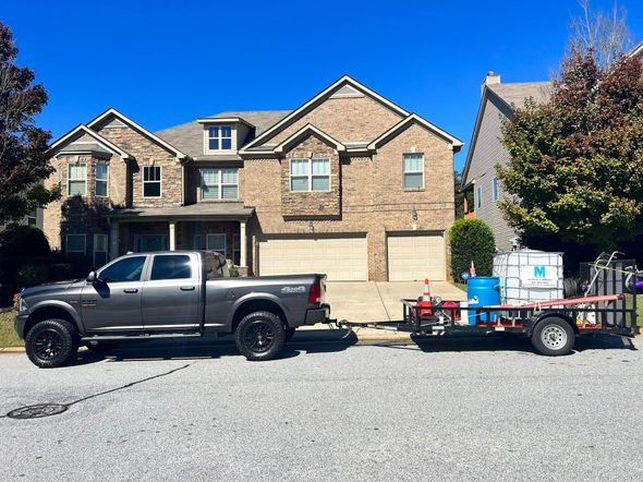 Grey truck towing trailer with equipment in front of a two-story brick house on a sunny day.