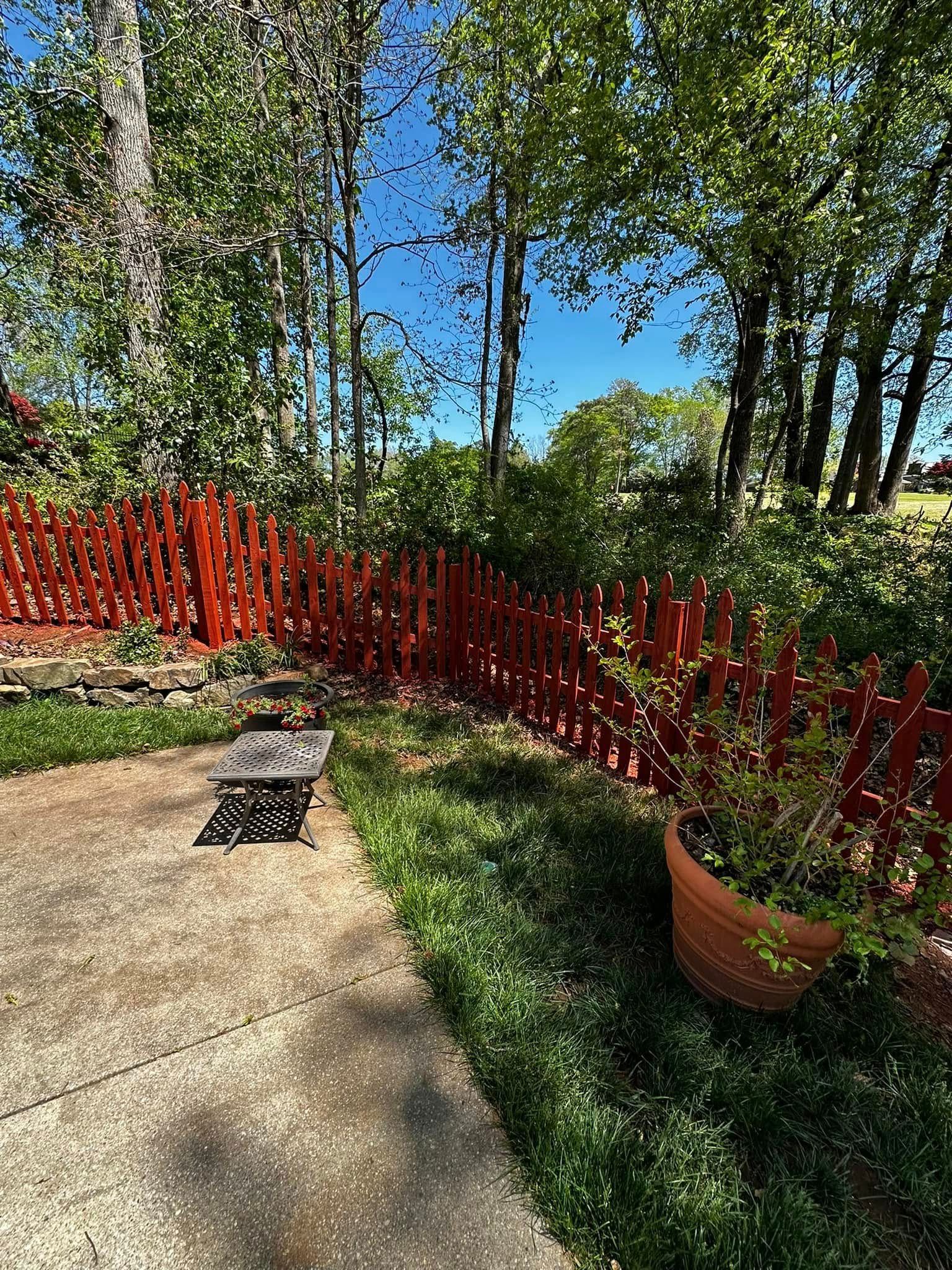Red picket fence borders a patio with greenery and trees on a sunny day.