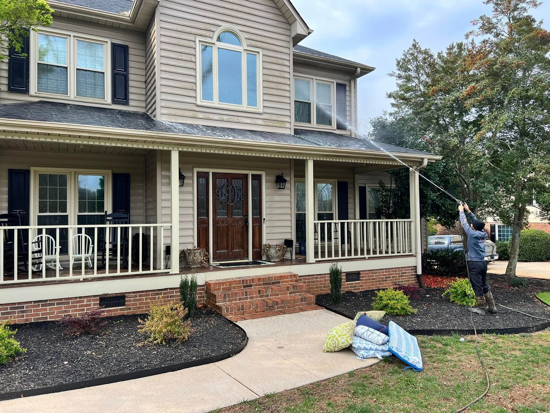 Person pressure washing a two-story house with beige siding. Front porch and landscaping in front.