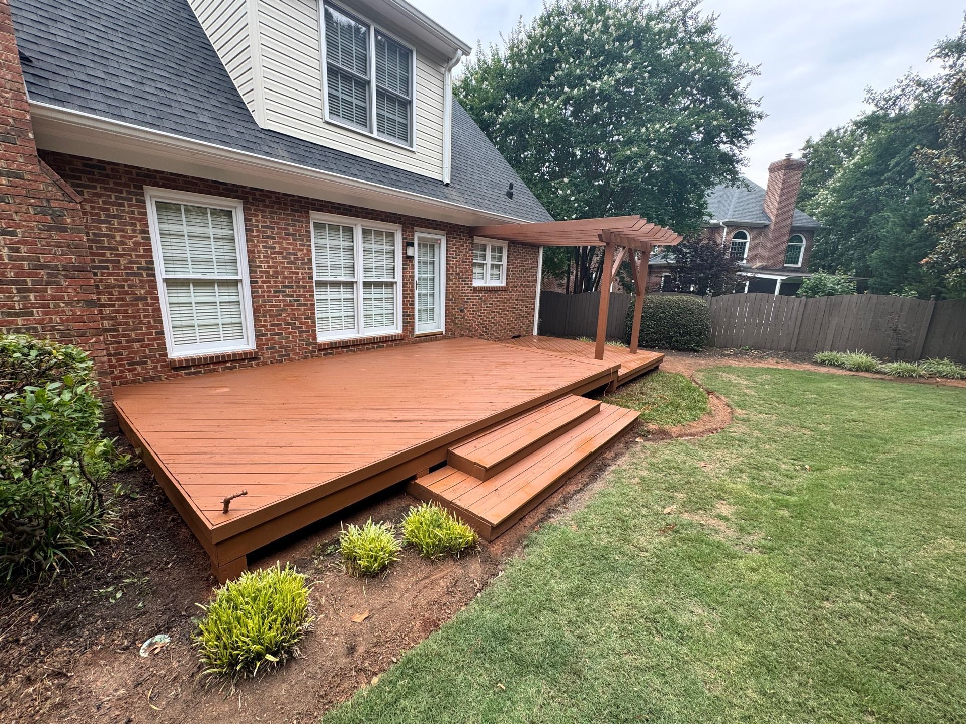 Red painted wooden deck with steps, pergola, and green lawn beside a brick house.