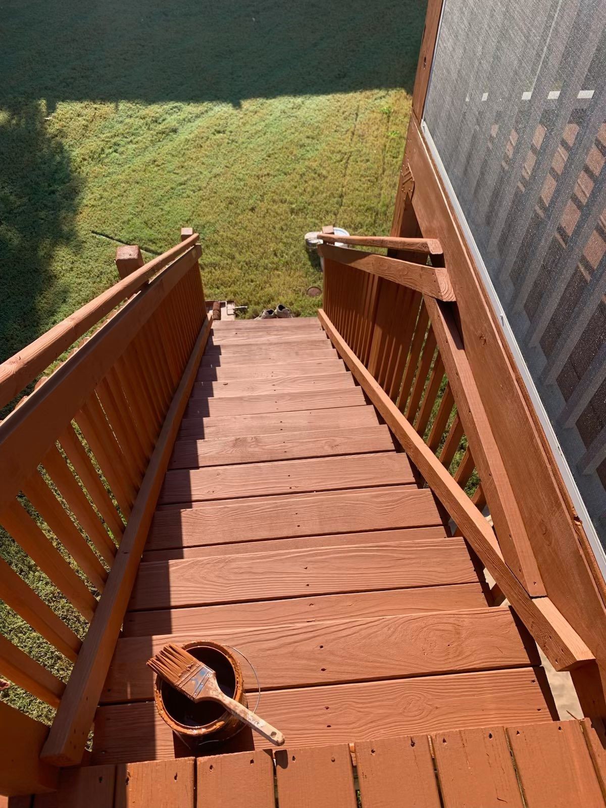 Wooden stairs painted reddish-brown, leading down to a grassy yard. A paintbrush rests on a paint can.