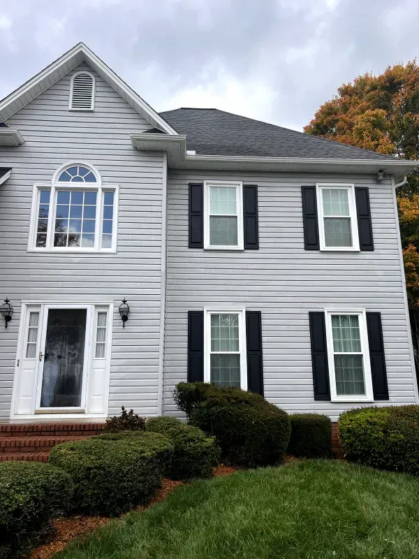 Two-story gray house with black shutters, arched window above the front door, and green bushes in front.