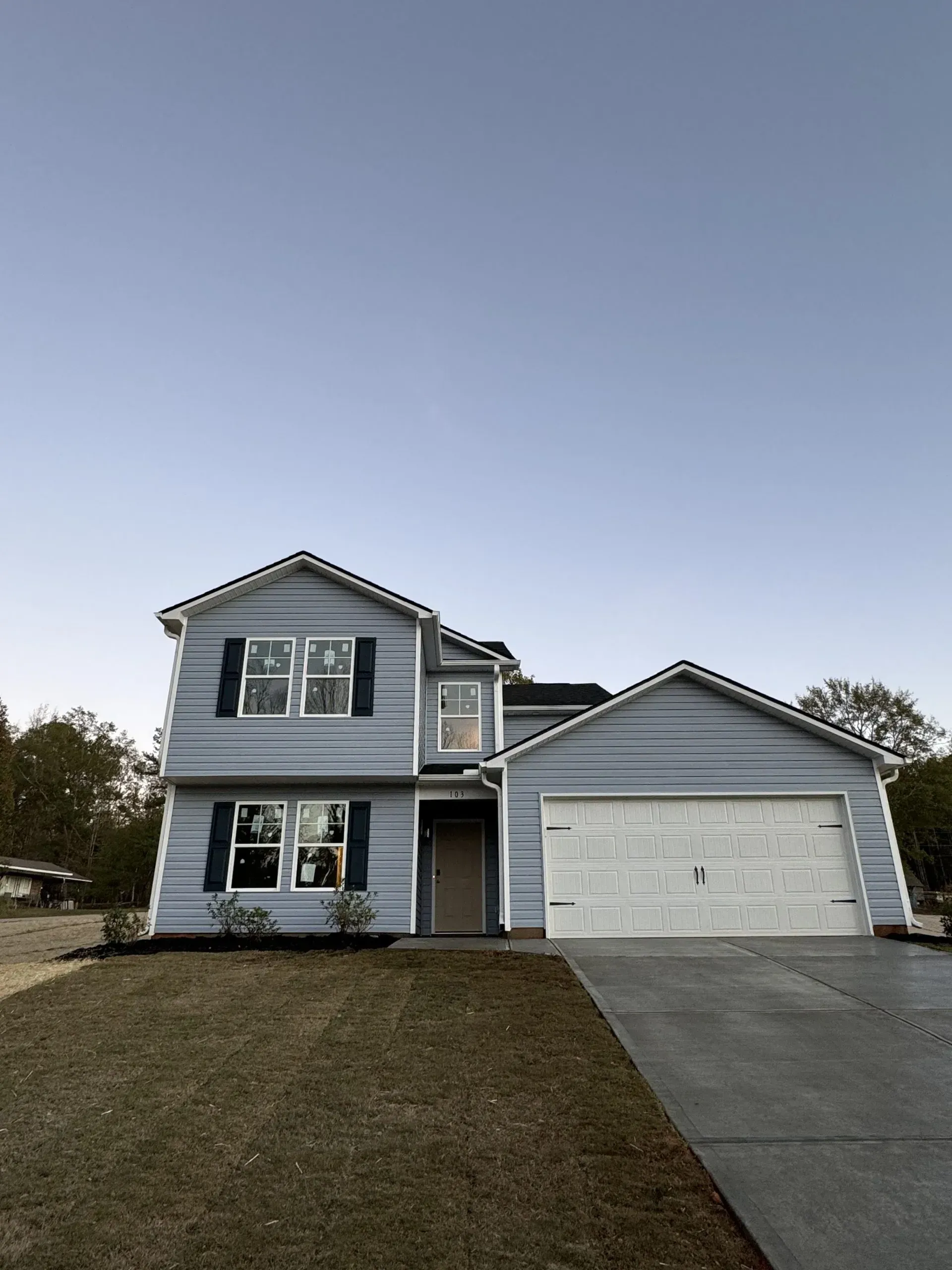 Two-story blue house with a garage and dark blue shutters, on a grassy lawn with a concrete driveway.