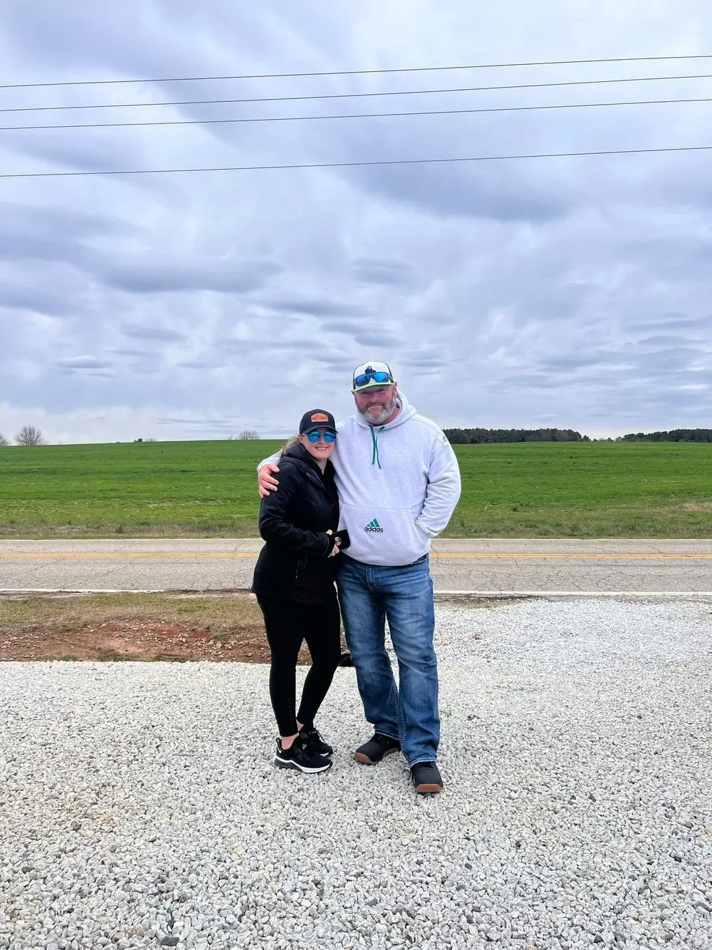 Couple standing outside, woman with arm around man; cloudy sky, green field in the background.