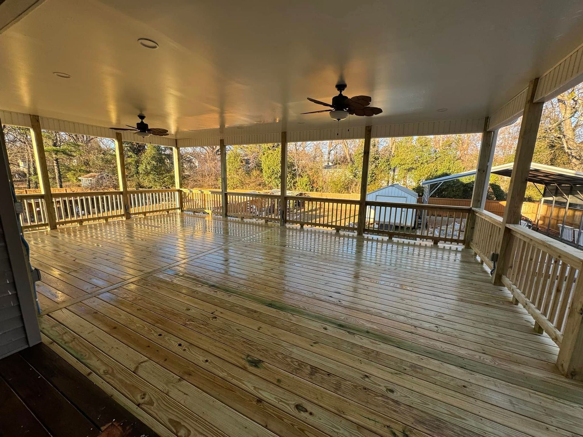 Covered wooden deck with a railing, overlooking a wooded area.