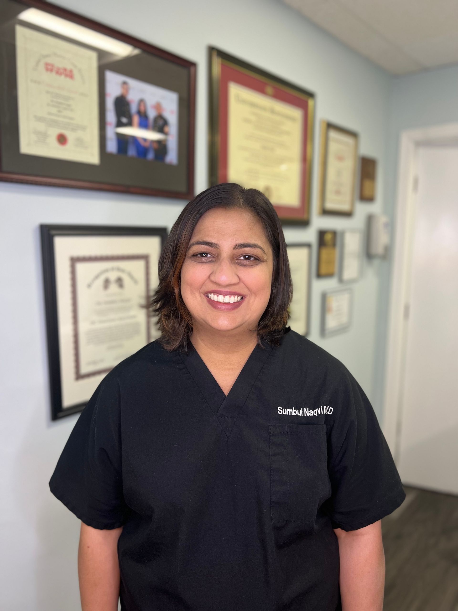 A woman in a black scrub top is smiling in front of a wall of framed pictures.