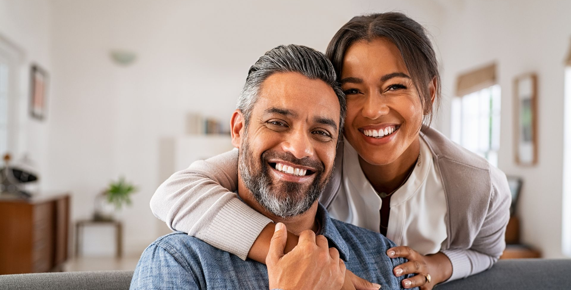 Smiling couple embracing in a well-lit living room; woman hugs man, both looking at the camera.