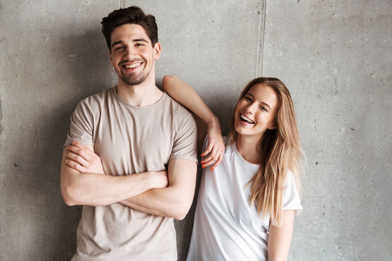 Man with arms crossed and woman smiling, leaning against a concrete wall. Both are smiling, wearing t-shirts.