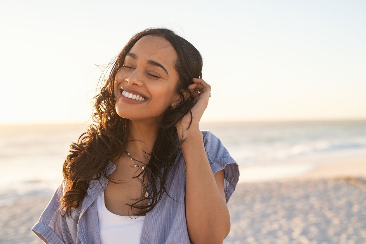 Woman smiling on beach, eyes closed, hand in hair, ocean in background, bathed in sunlight.