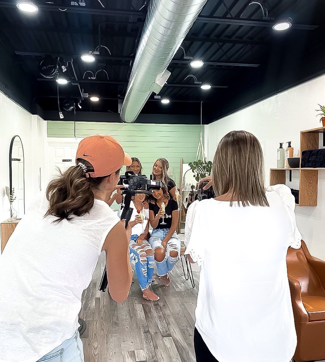 Two people taking photos of others at a salon, brightly lit, with styling chairs and a mirrored wall.