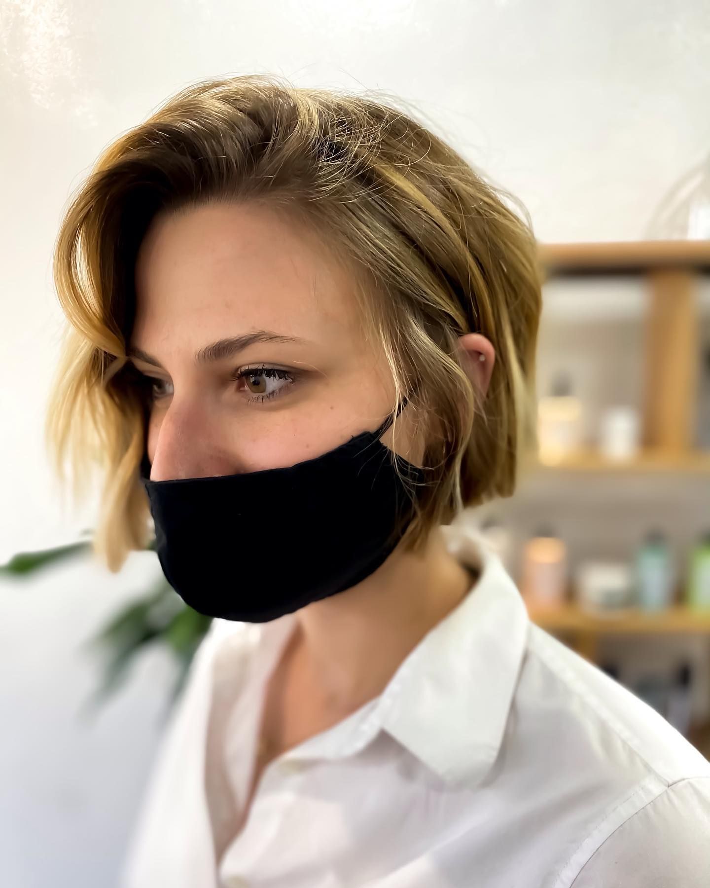 Woman with short, layered blonde hair wearing a black face mask and white button-down shirt, in a salon.