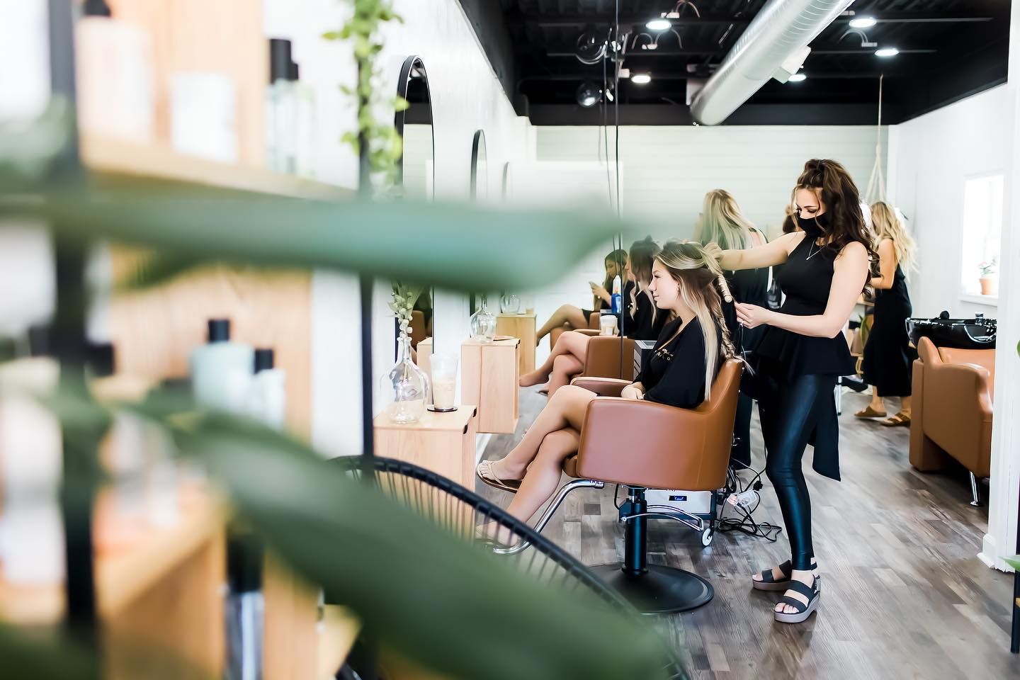 Hair salon: stylist styling client's hair in front of a mirror. Modern interior, bright lighting, other clients present.