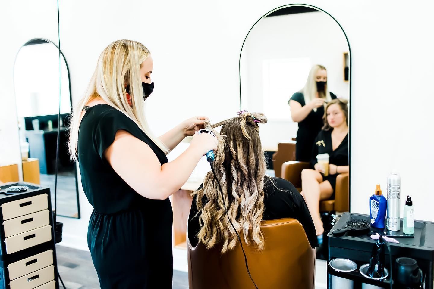 Hair stylist curling a client's hair in a salon; another stylist in the background.