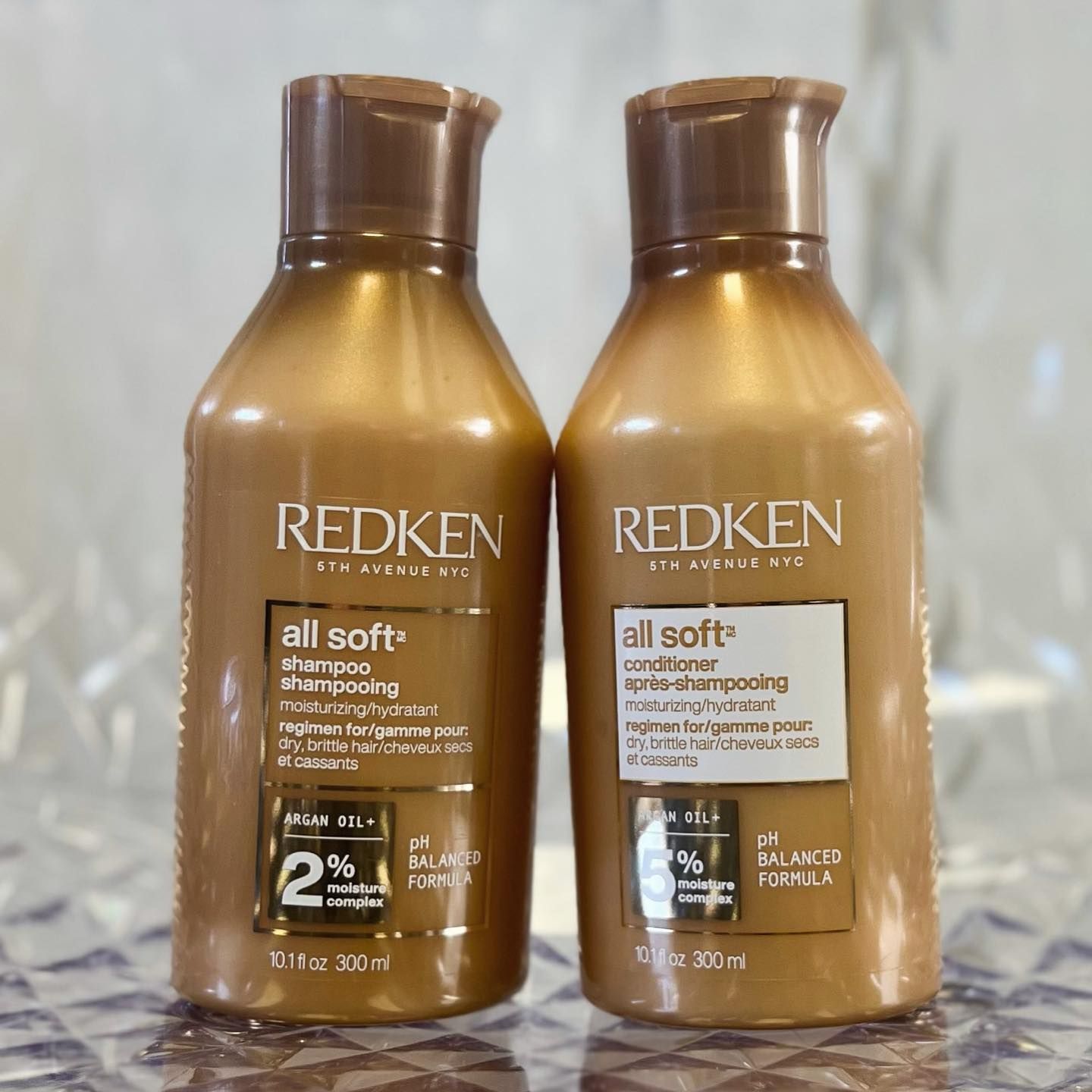 Two Redken All Soft hair product bottles. Gold-colored bottles with brown lids. Shampoo and conditioner are shown.