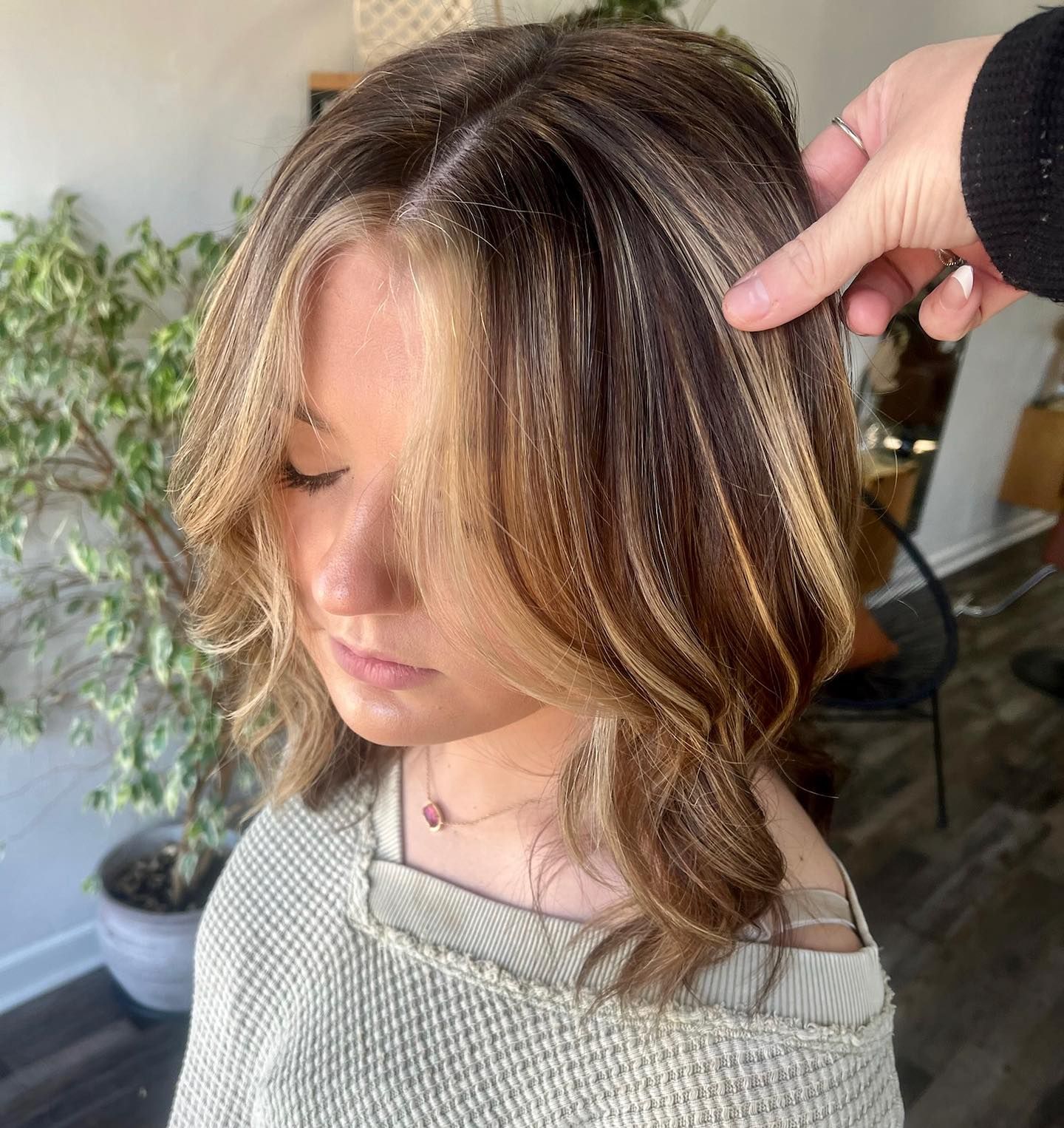 Woman with highlighted hair, hand touching hair; indoors with potted plant.