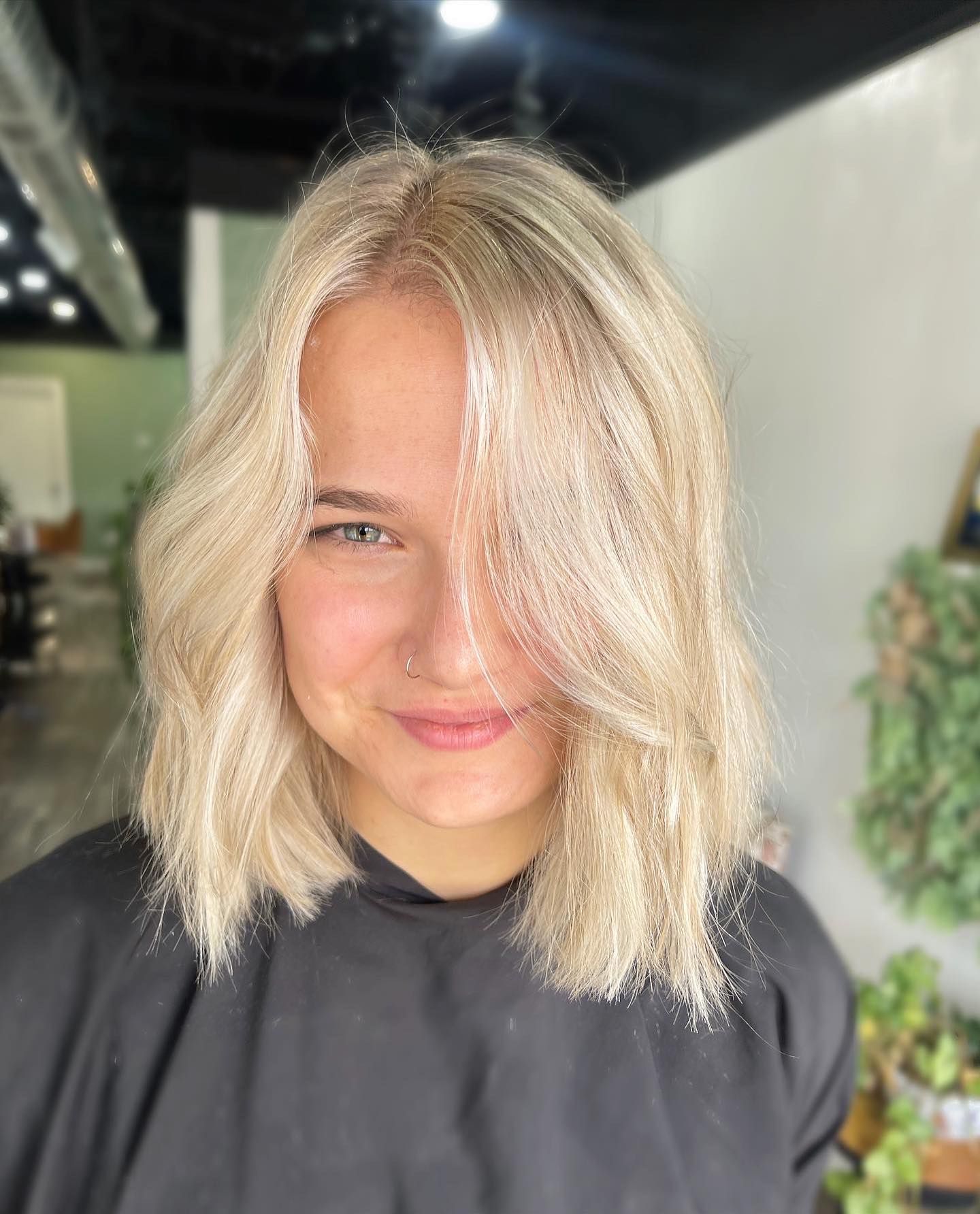 Blonde-haired person with wavy hair, smiling.  Wearing black salon cape. Indoors with green wall and plants.