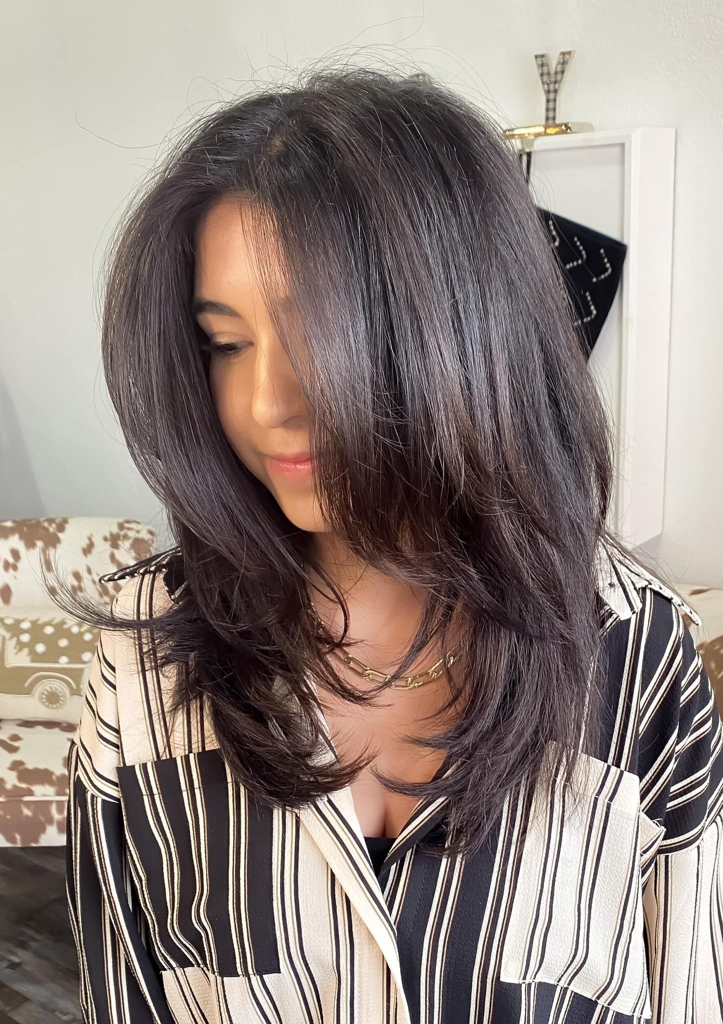 Woman with long dark layered hair, wearing a patterned shirt, looking down.