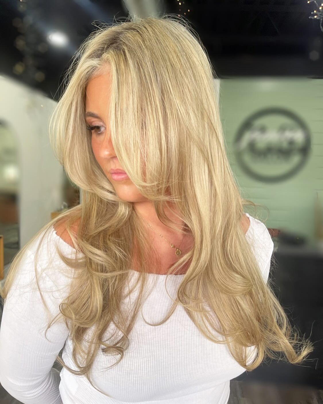 Woman with crimped blonde hair, layered around face, wearing white off-shoulder top.