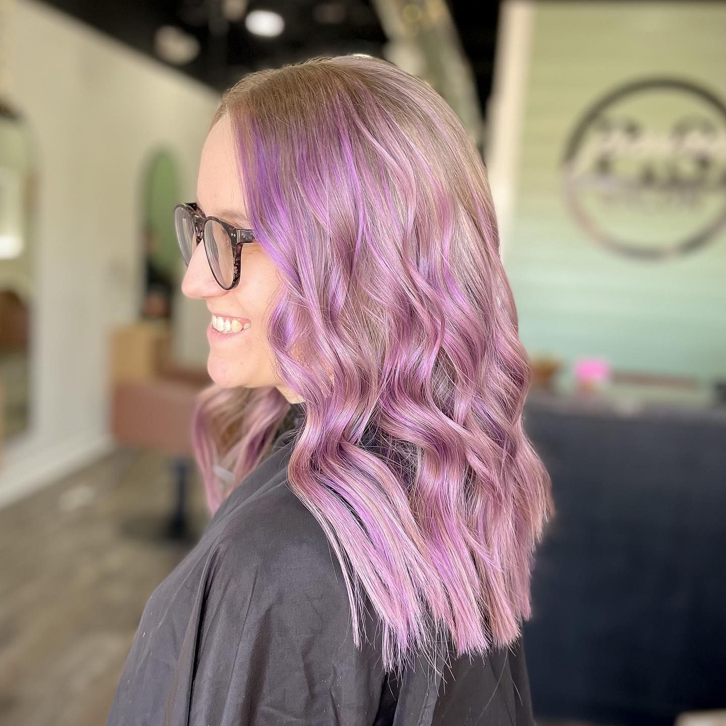 Woman with purple wavy hair and glasses smiles in a salon.