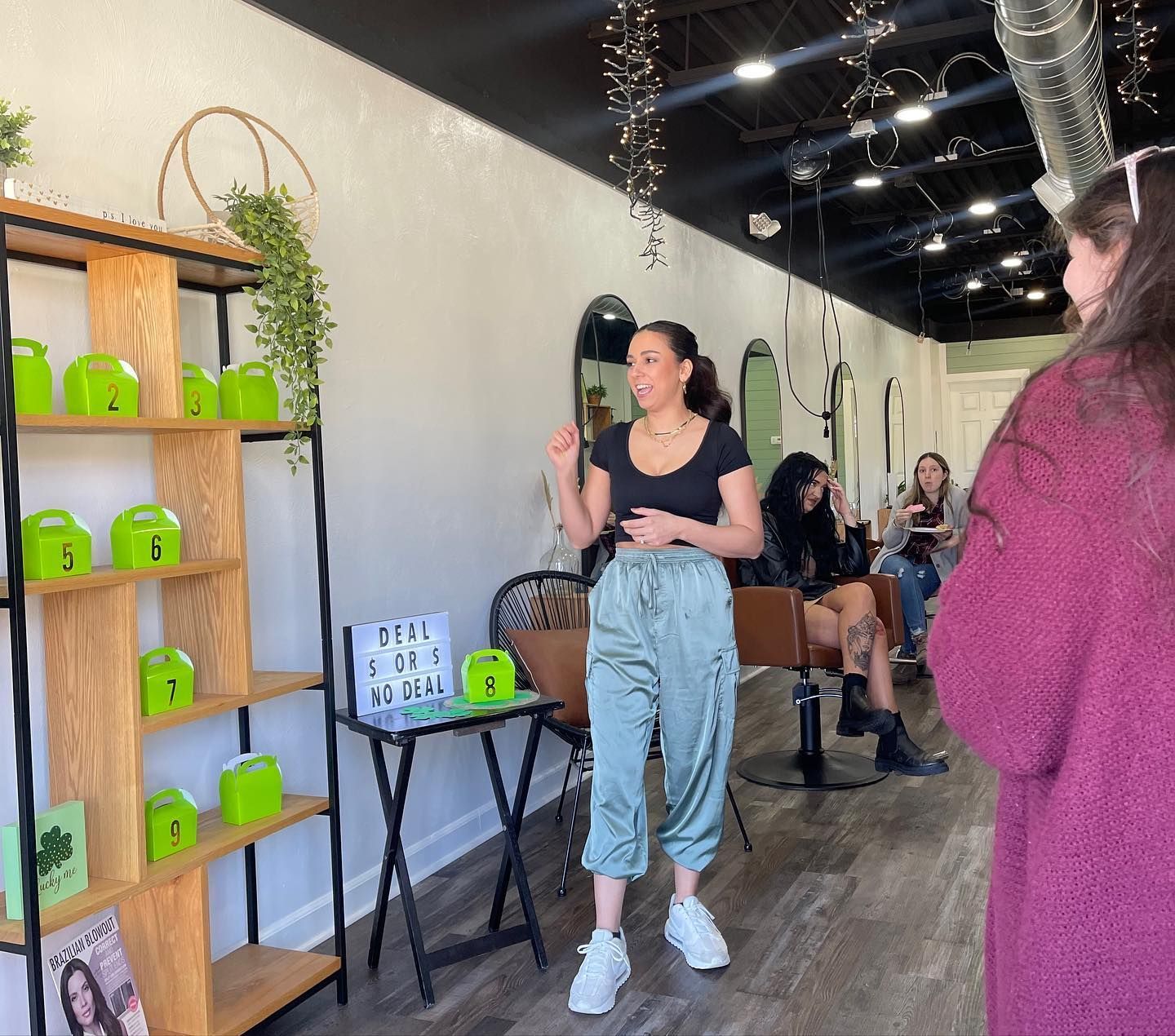 Woman in salon gestures, facing an audience. Green decorations and mirrors on the wall.