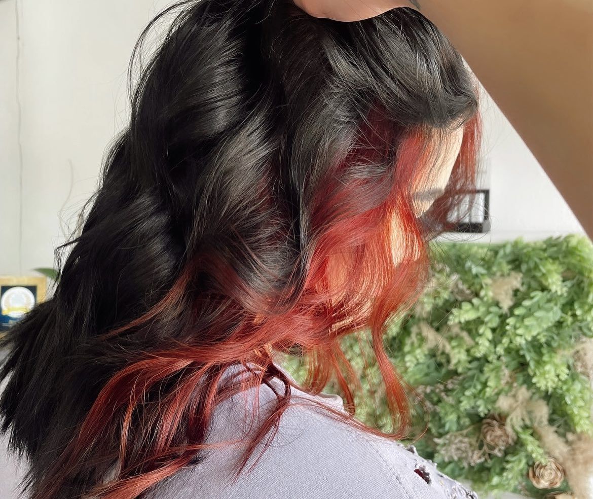 Woman with black hair, red highlights, and wavy style, with a white wall and green plant in background.
