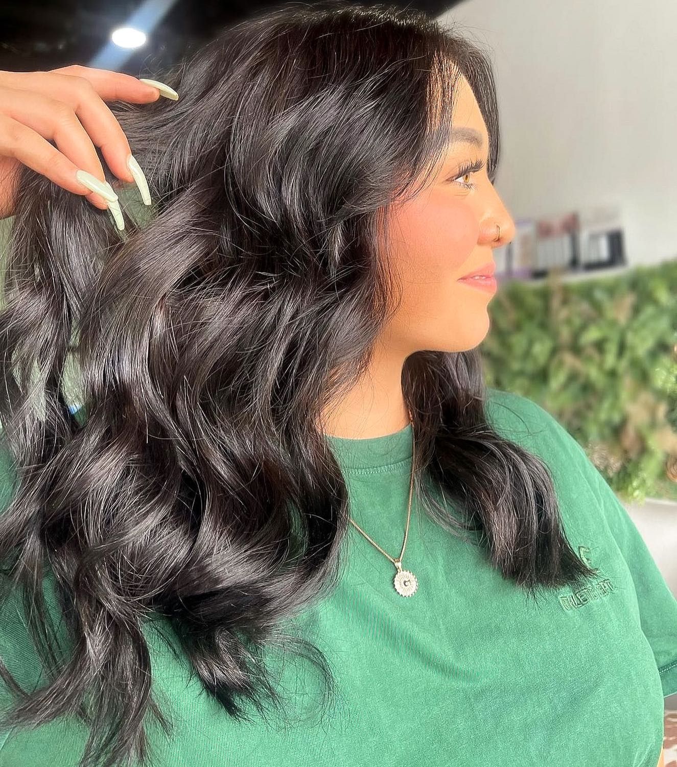 Woman with long, wavy black hair, wearing a green sweater and necklace, touches her hair.
