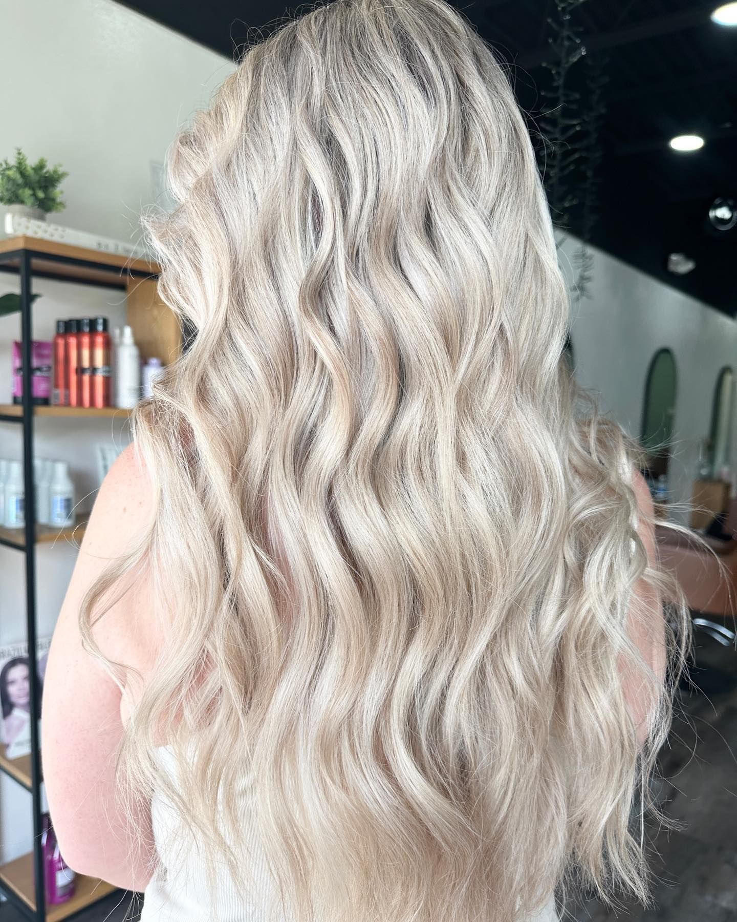 Woman with long, wavy, light blonde hair.  In a salon, with products in the background.