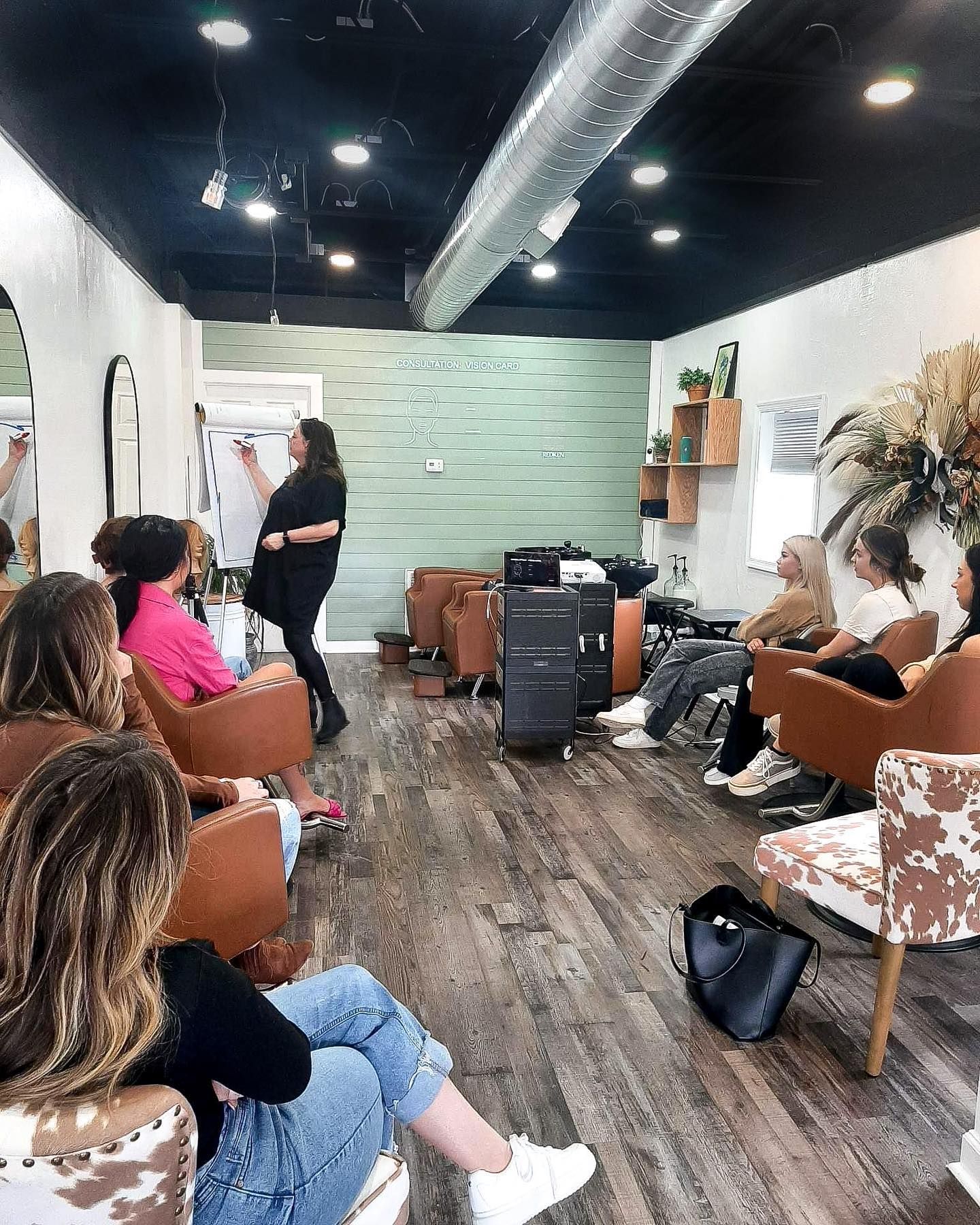 A woman teaching a class in a hair salon. Students are seated in brown and cowhide chairs.