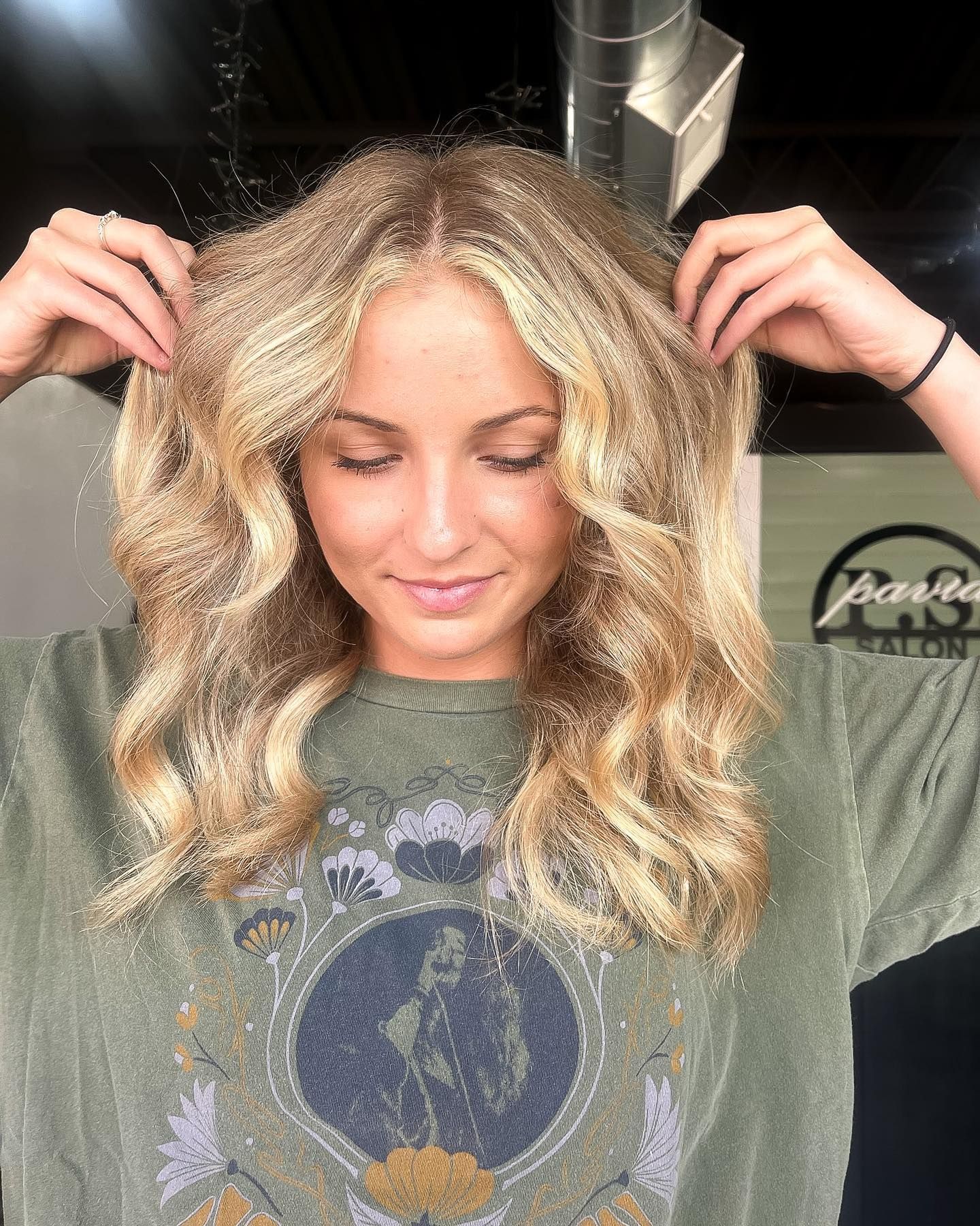 Woman with wavy blonde hair, wearing a green graphic t-shirt, touching her hair with a salon background.