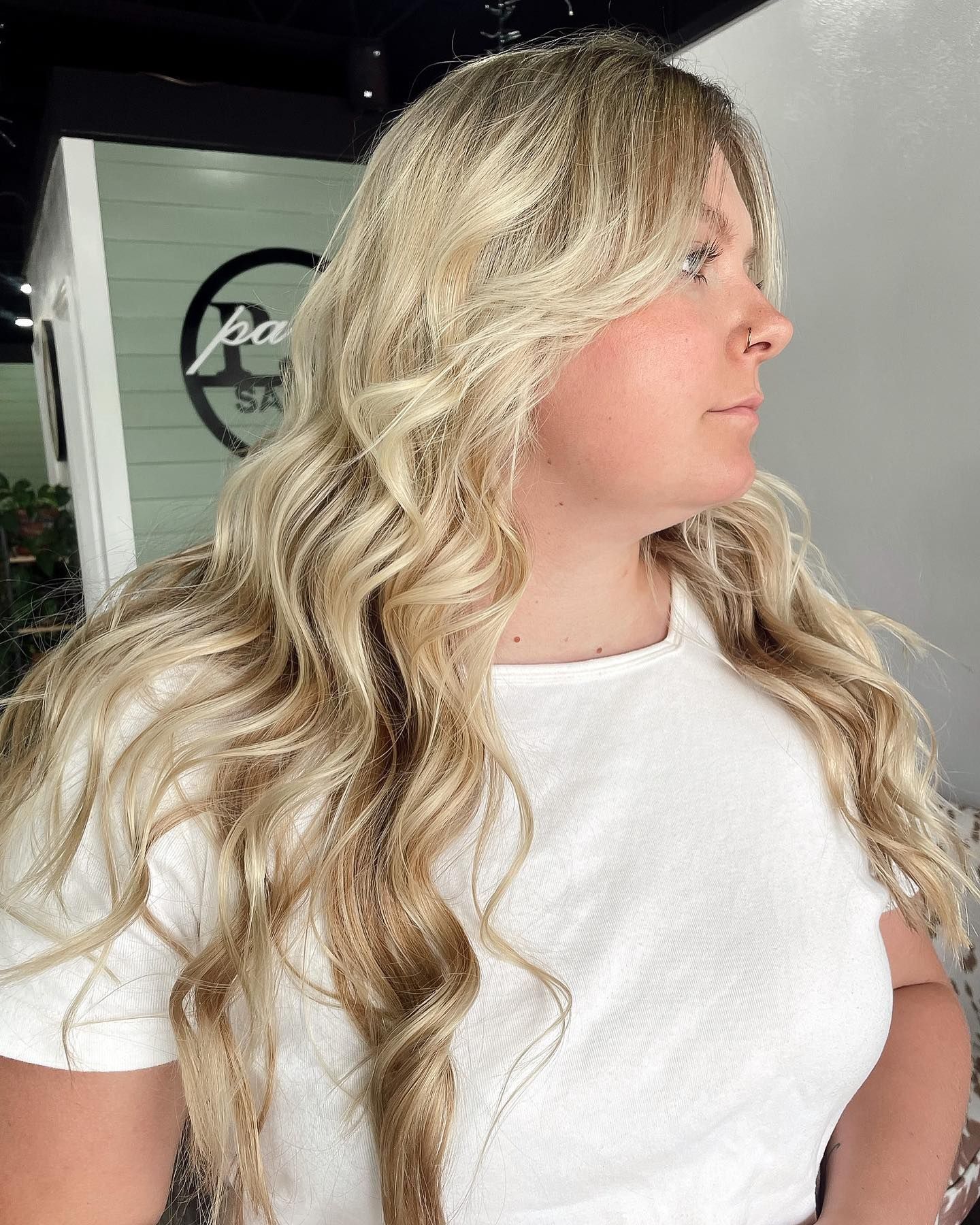 Blonde woman with long wavy hair, in a white shirt, looking to the side. Salon background.
