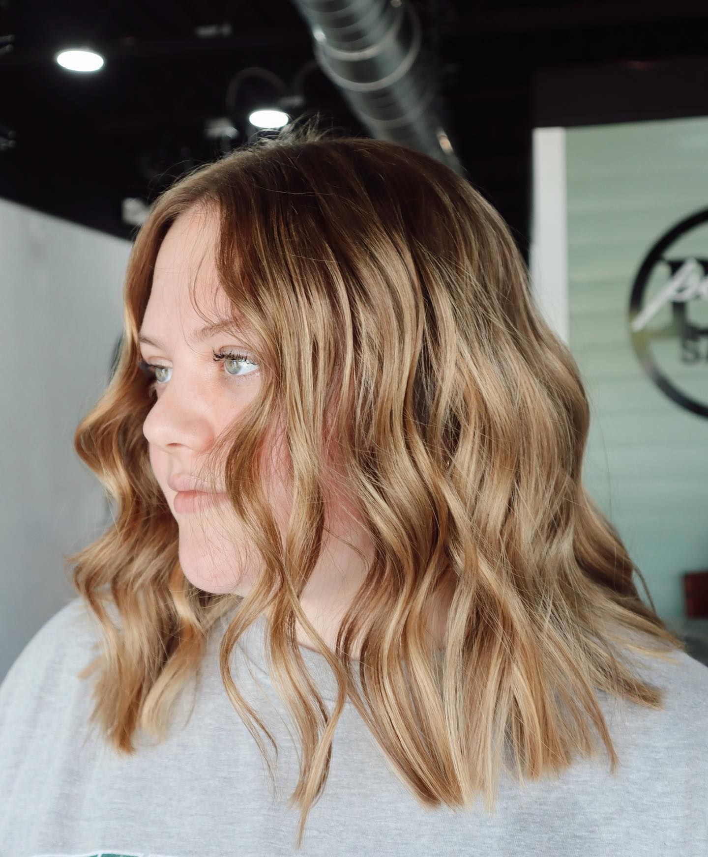 Woman with wavy, blonde-highlighted hair, in a salon setting, looking to the left.