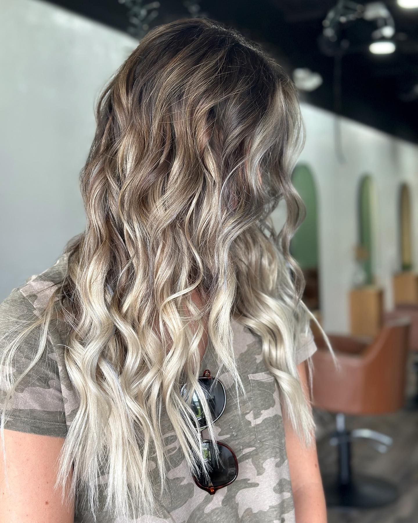 Woman with wavy, blonde-highlighted hair wearing a camouflage shirt in a salon.